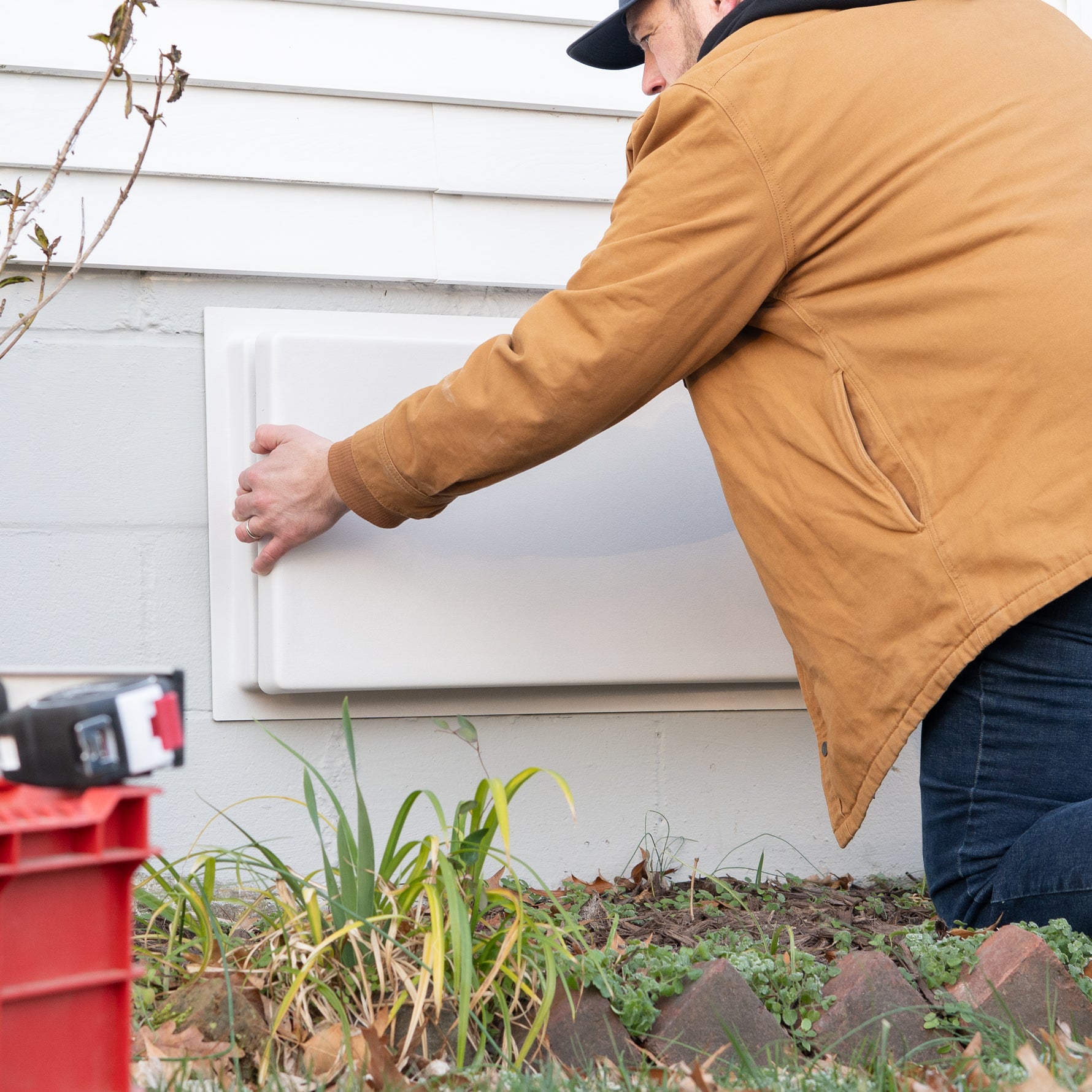 Person in a brown coat opening an outdoor white crawl space door cover on a house exterior.