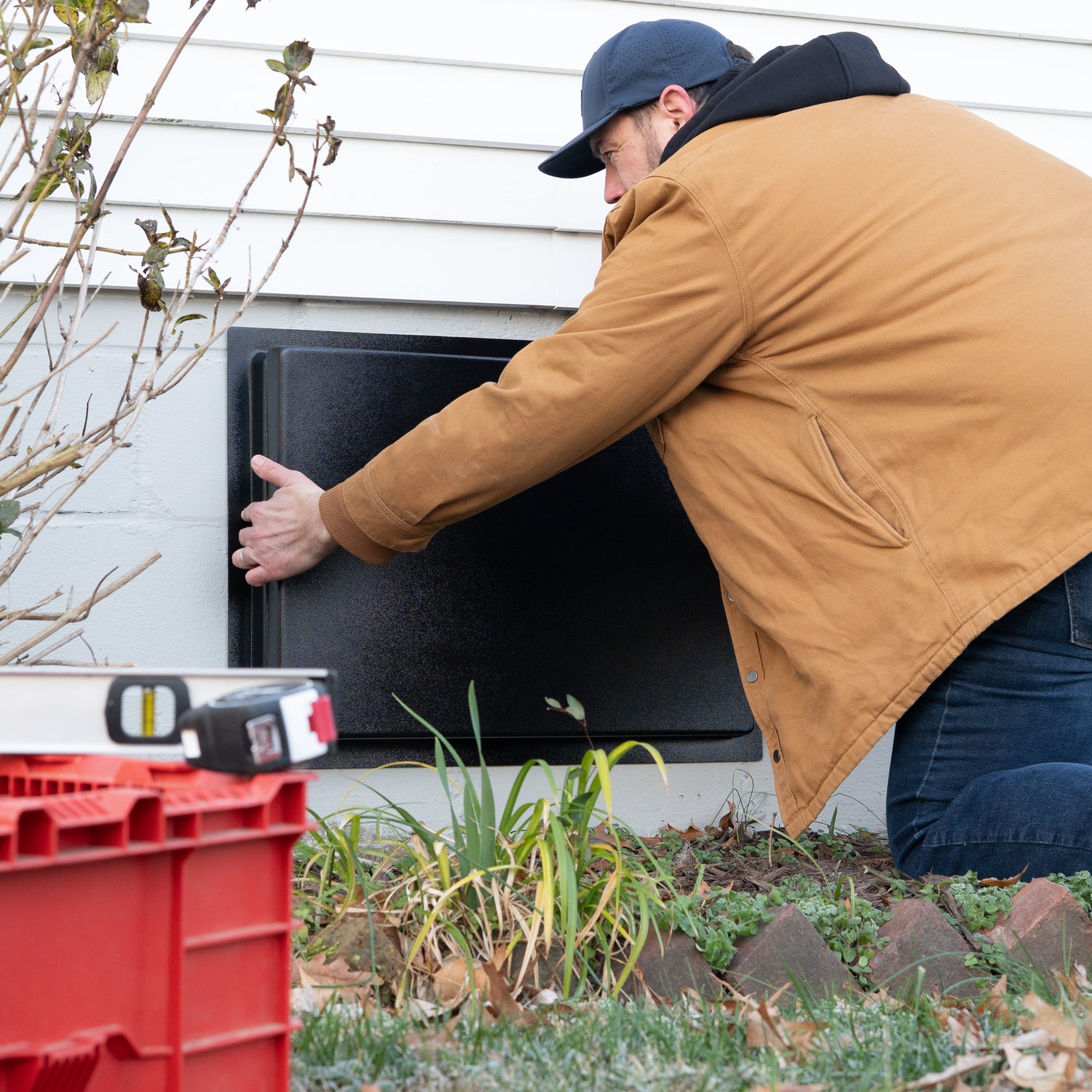 Person opening a black crawl space door with a red toolbox in the foreground.
