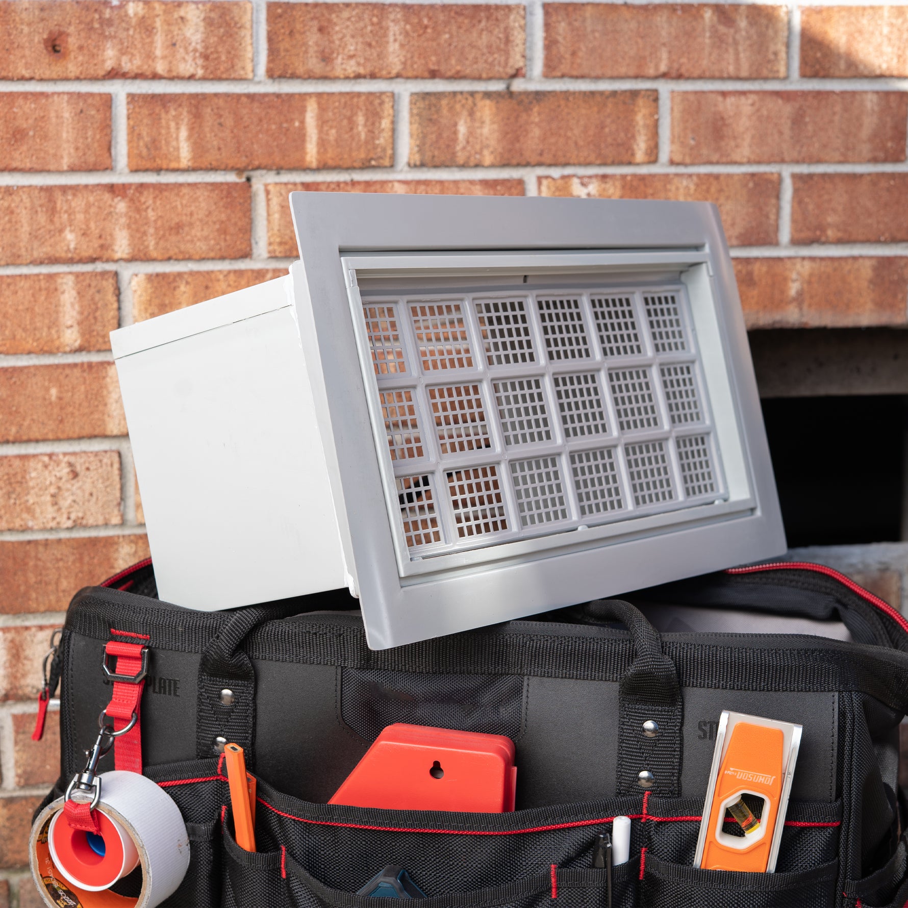 Tool bag with tools and a gray flood vent against a brick wall 