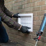 Person installing a white rectangular vent cover on a brick wall