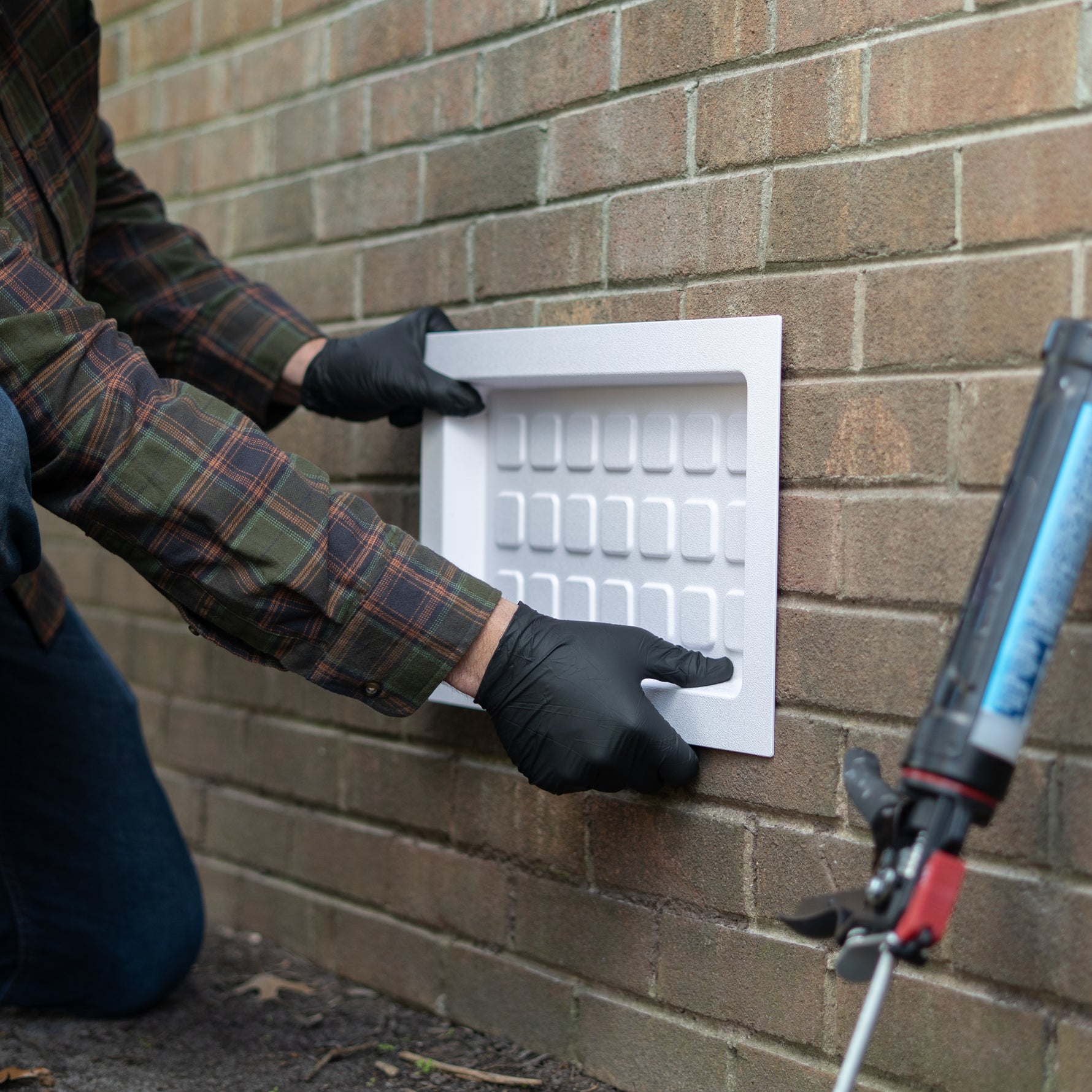 Person installing a white rectangular vent cover on a brick wall