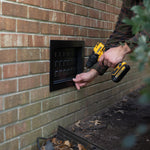 Person using a drill to install a black recessed vent cover on a brick wall.