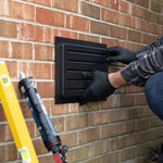 Person installing a black outward mounted outdoor vent cover on a brick wall with tools nearby.